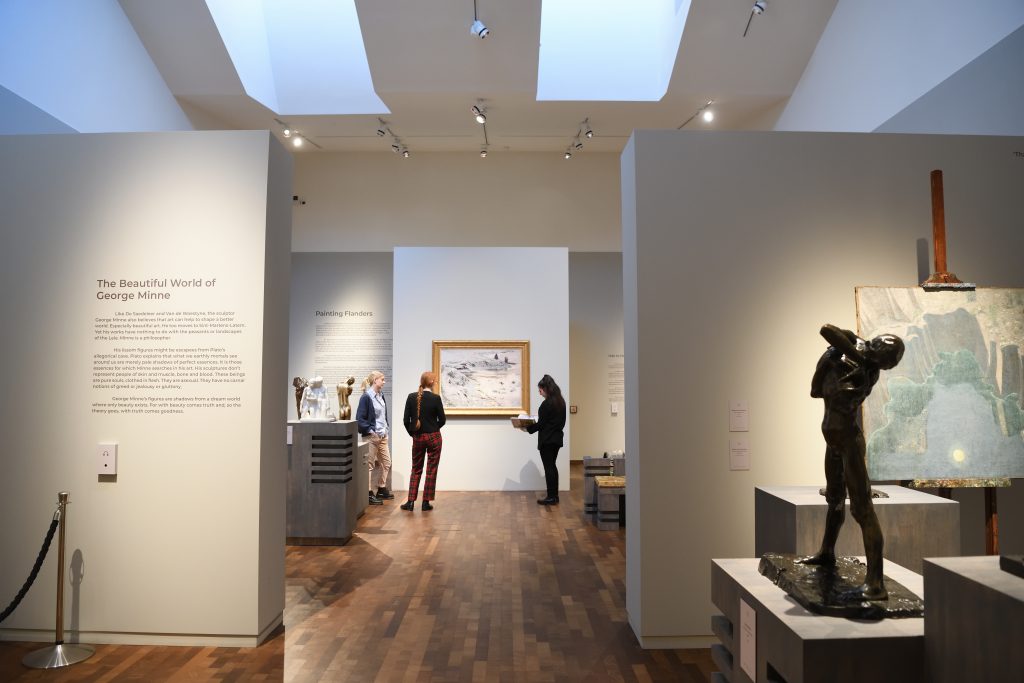 An image taken in the Timothy and Mary Clode Gallery at Gainsborough's House. There are various artworks and three people viewing and discussing the work.