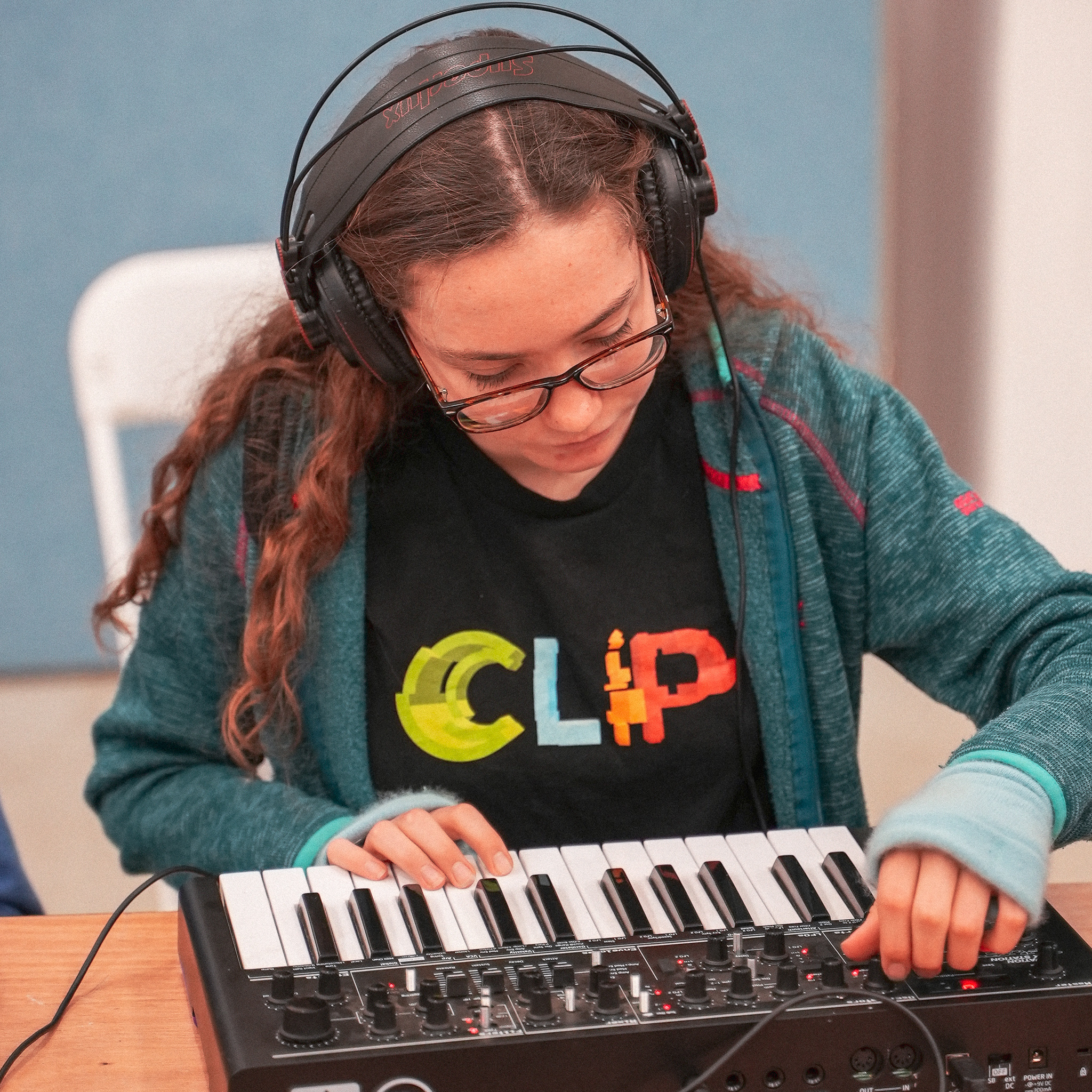 An image of a young person playing with a keyboard. They have headphones on and a t-shirt on with the CLIP logo on it.