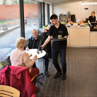 An image taken within The Watering Place café. There are various tables and chairs and a few visitors being served by café staff member, Mitch.