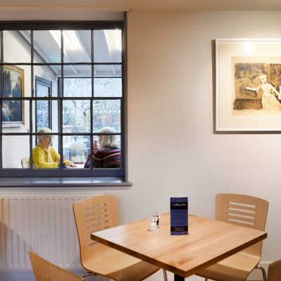An image taken within The Watering Place café at Gainsborough's House. It depicts two visitors sat at a table chatting over coffee.