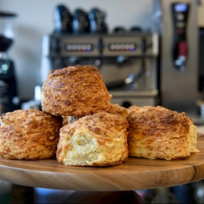 A close-up image of freshly-baked cheese scones on a wooden platter. There are coffee machines in the background.