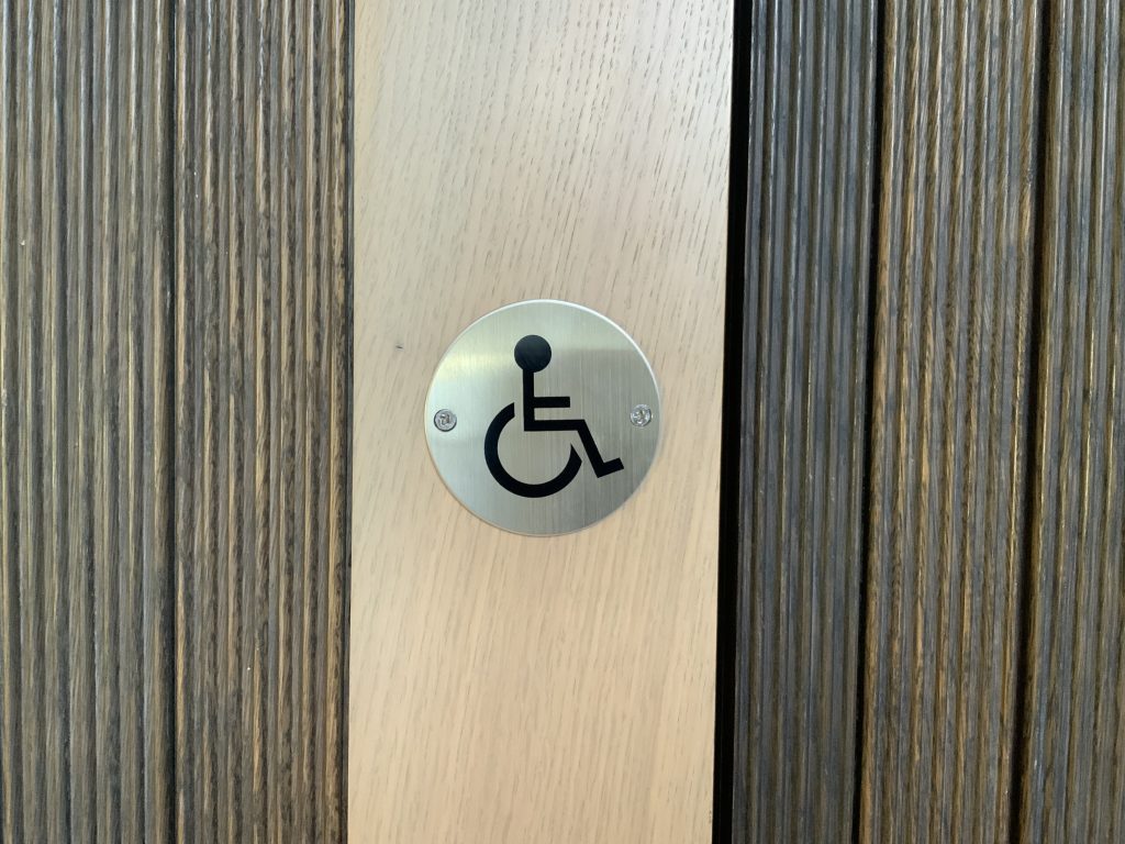 An image taken outside one of Gainsborough's House disabled toilets. There is wood detail with a metal plaque with the disabled icon on it.
