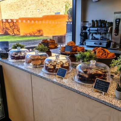 An image taken of the pastry counter in The Watering Place café. It depicts a range of cakes, pastries and sweet treats. There is a coffee machine in the background and the walled garden flow effortlessly in through the large windows.