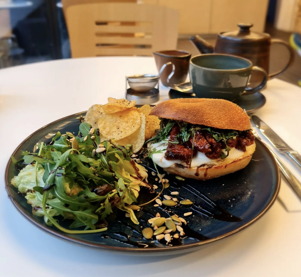 An image of a toasted bagel on a plate with salad and crisps. There is a tea set in the background.