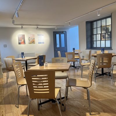An image taken within The Watering Place café. It shows the seating arrangement in room 1 of the café. There are exhibition posters in the background along with various tables and chairs for individuals and larger groups or families.