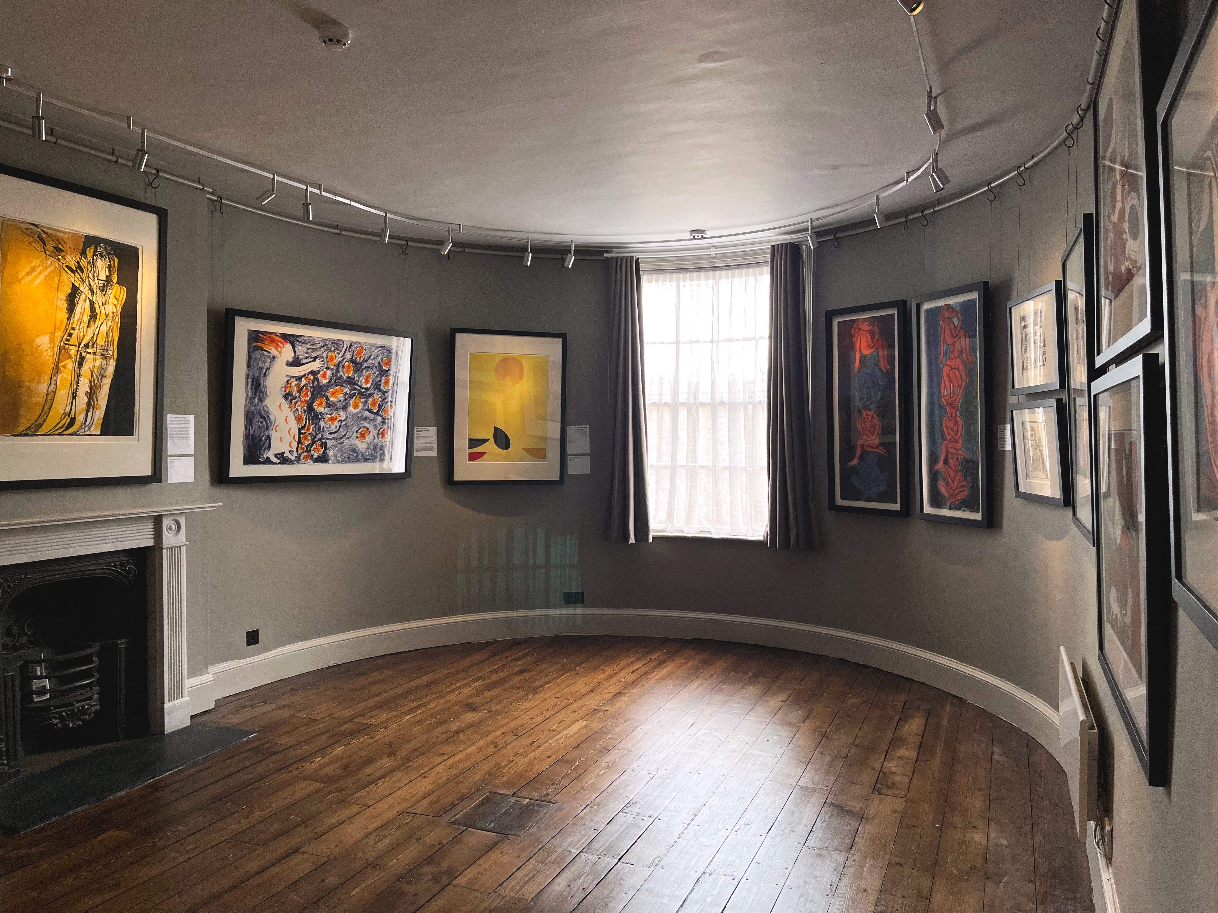 An image taken of the Striking Editions lithograph exhibition. There are numerous prints framed on the walls. The Upper Bow Room has a rounded back wall.