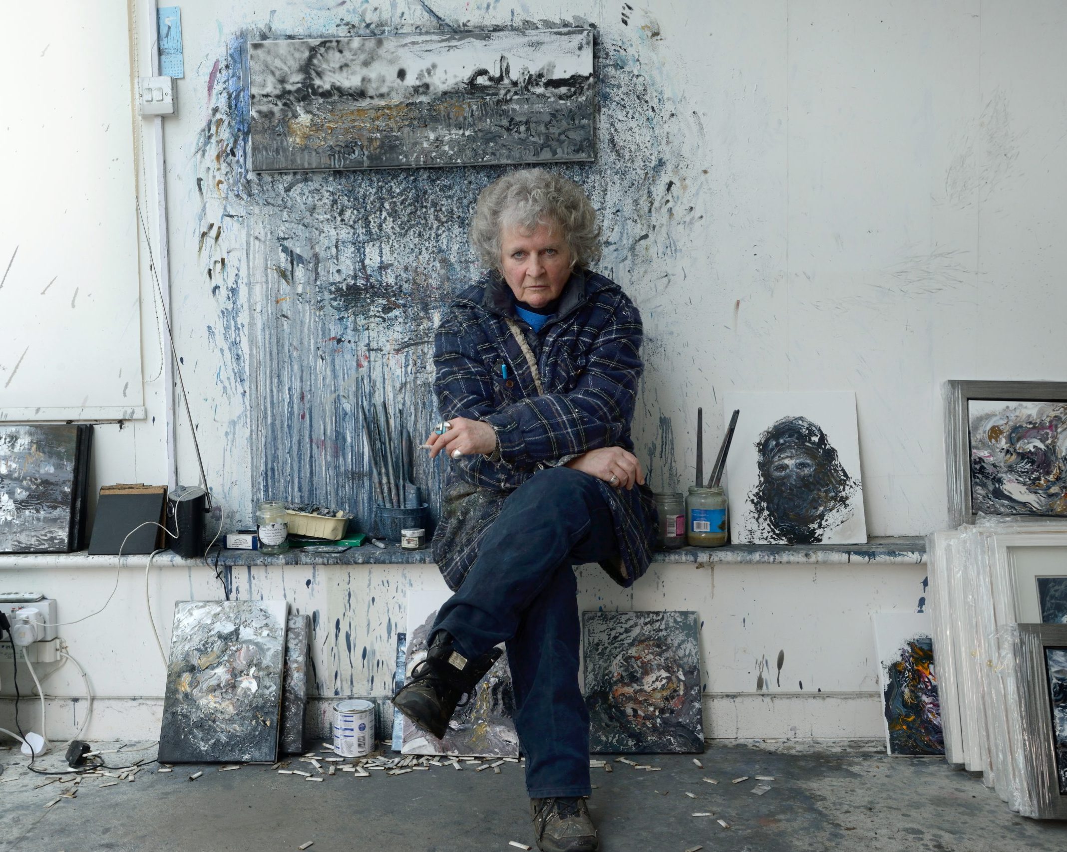 An image of the artist in their studio, Maggi Hambling. She has numerous cigarette butts around her while holding a lit cigarette in hand. Her expression is moody and stern.