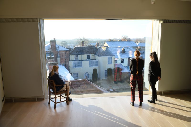 An image of three people looking out onto Gainsborough's House. They are stood and sat in the Landscape Gallery with sun shining through the large window.