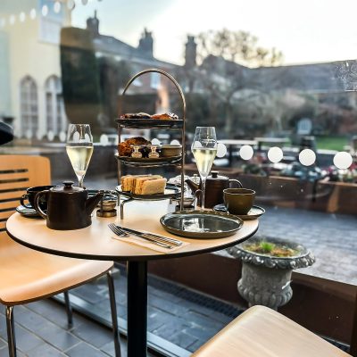 An image taken of an afternoon tea inside The Watering Place café, overlooking Gainsborough's house walled-gardens. There are various baked goods on a 3-tier display. There is also tea and Prosecco on the table.