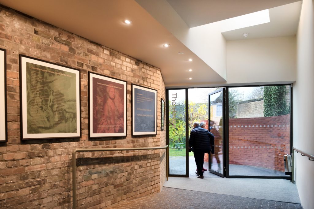 An image taken in the lobby at Gainsborough's House. There are 4 framed silk displays hung on a brick wall to the left. There ar etwo people exiting through a larg eglass door to the right.