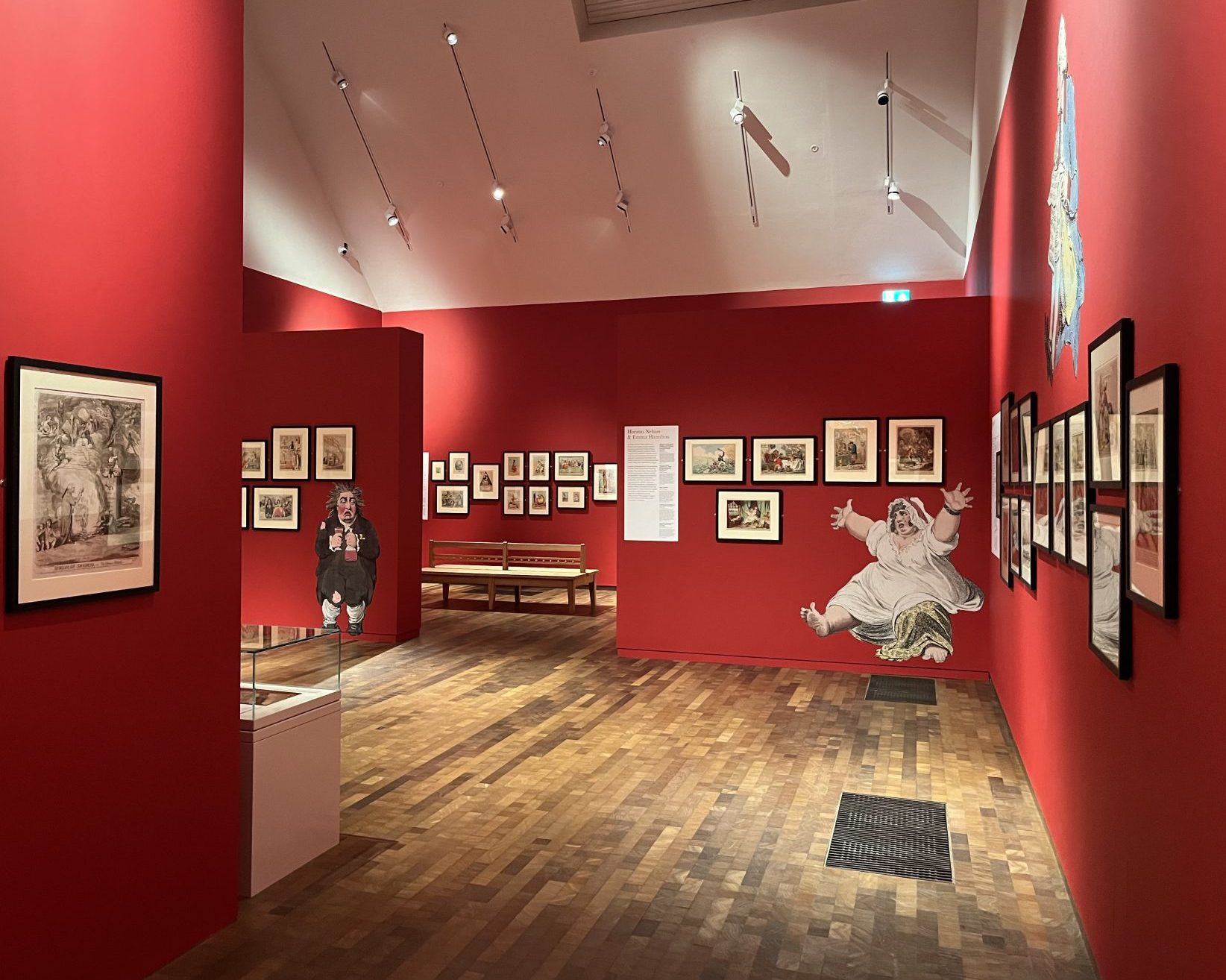 An image taken in the Timothy & Mary Clode Gallery at Gainsborough's House. The exhibition, James Gillray: Characters in Caricature can be seen There are multiple framed prints on red walls with large-scale character cut-outs placed beside them.