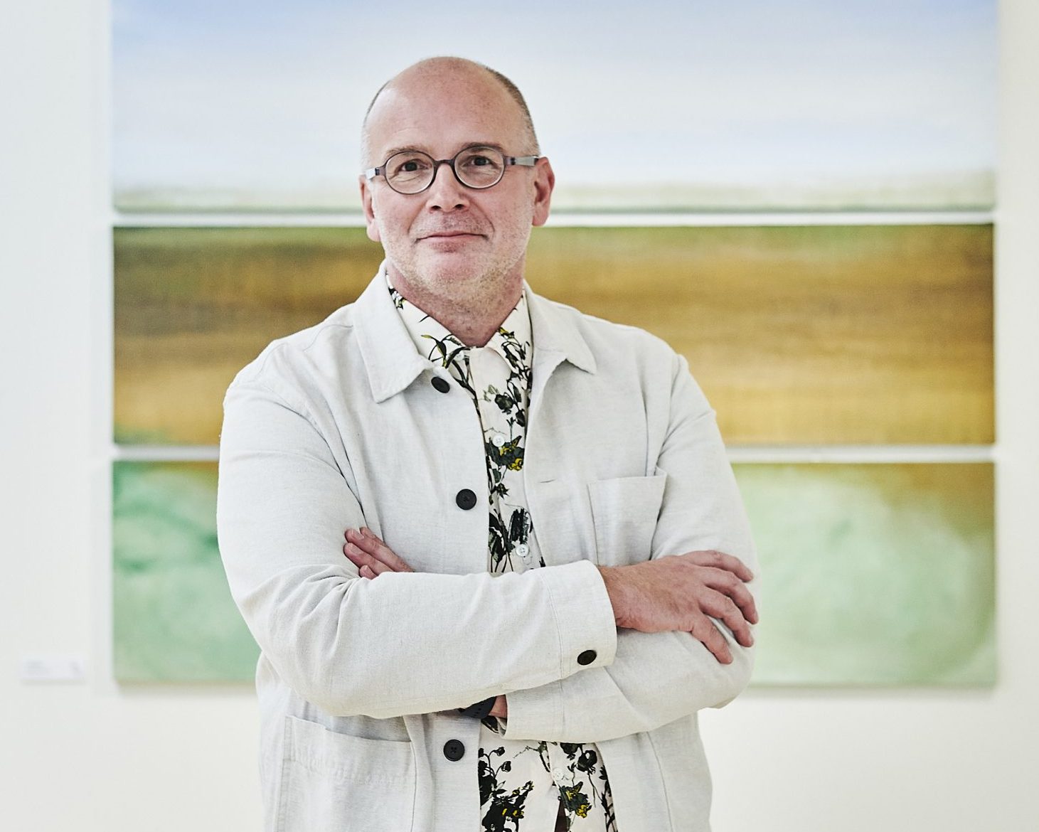 Simon Ofield Kerr is in main focus. He is wearing a cream jacket and floral shirt. His arms are crossed and he is smiling. There is an abstract painting behind him in the background.