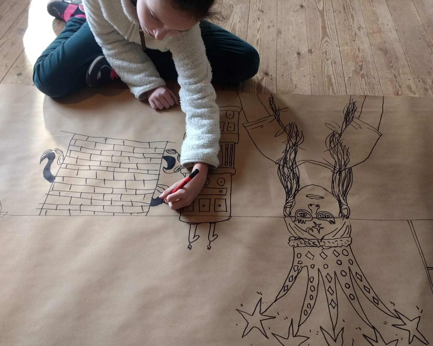 An image of a child drawing on brown paper while sat cross-legged on the floor.