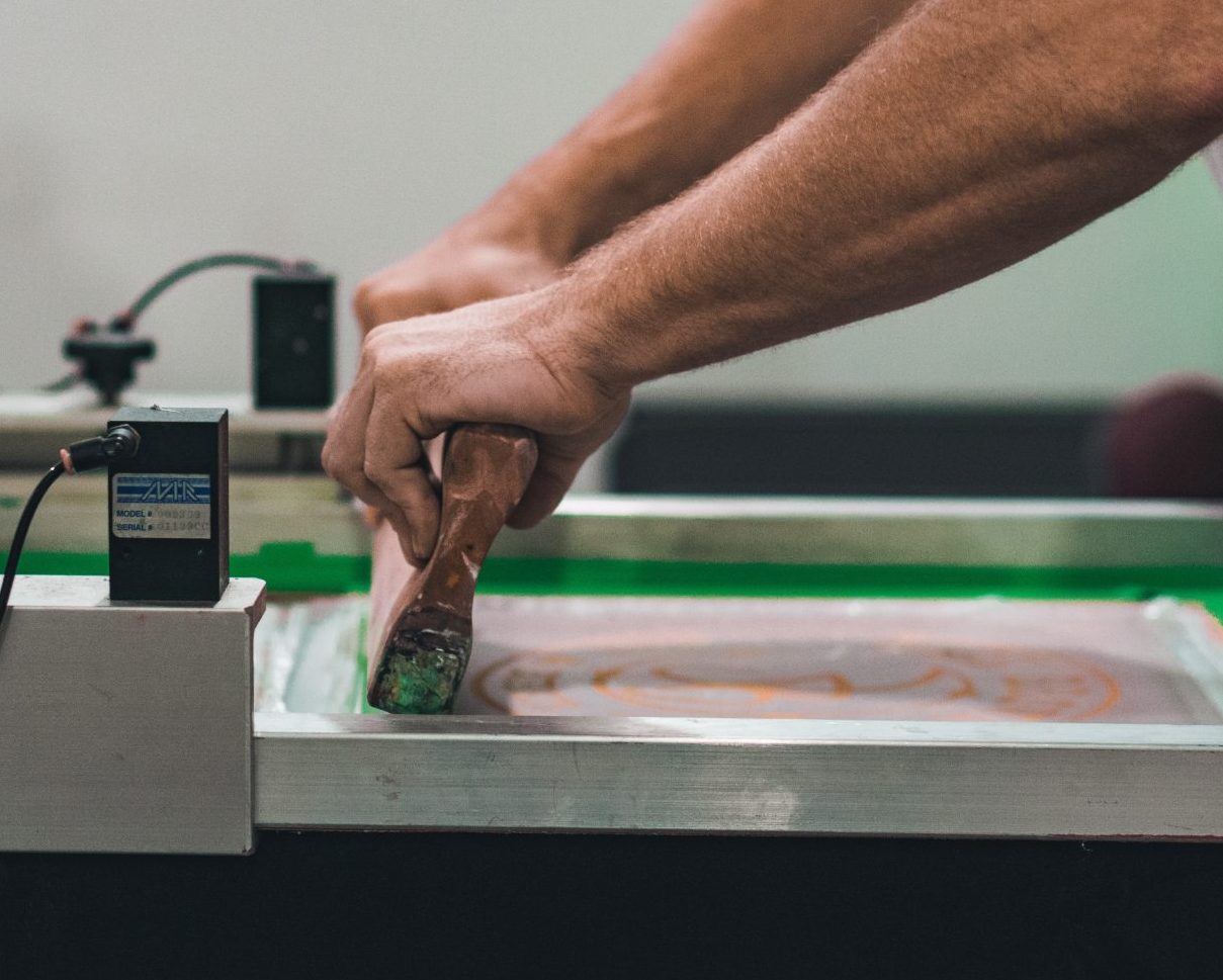 An image of a person using a screenprinting press. They are using a squeedgee on a t-shirt.