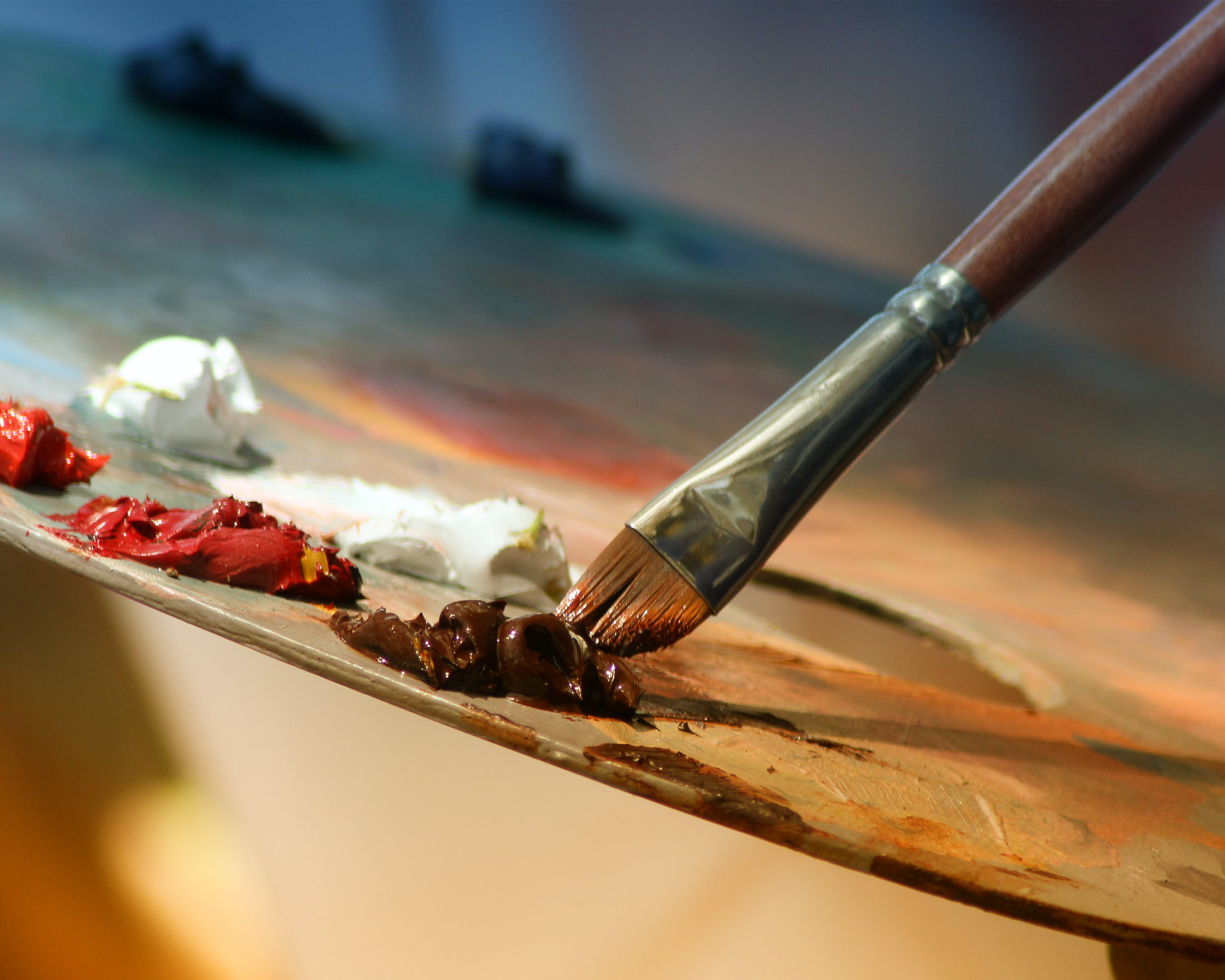 An image of a paintbrush picking up brown oil paint from a traditional paint palette.