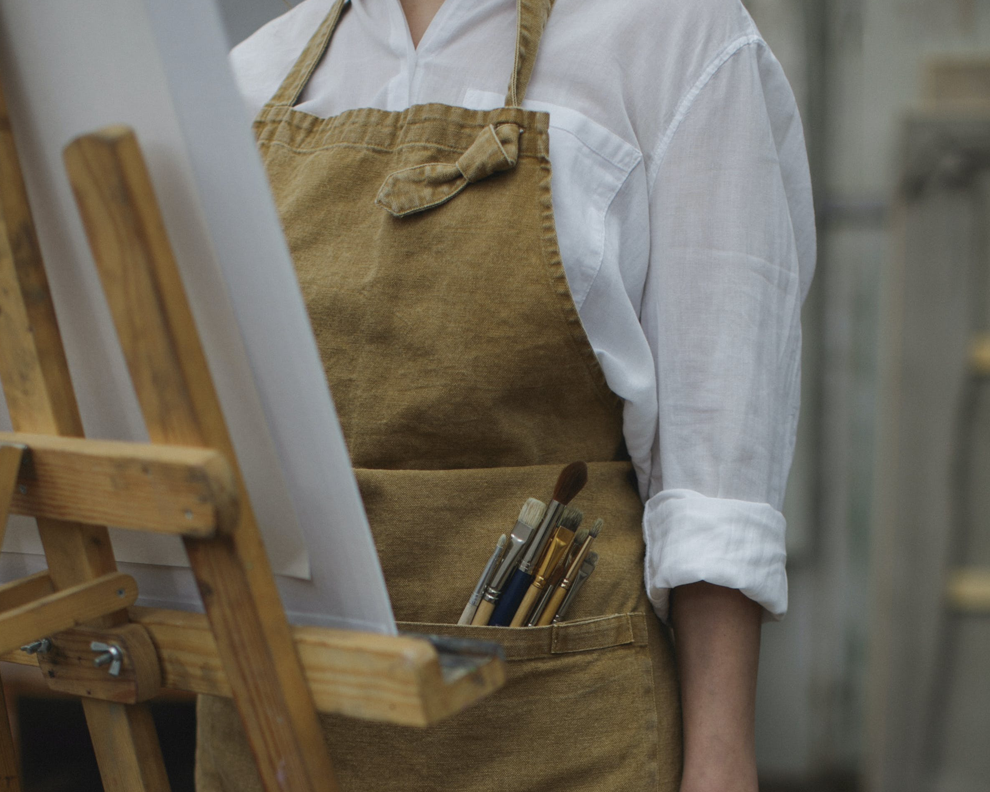 An image of a person stood in front of an easel with an apron on. They are wearing a white shirt and have paint brushes sticking out of their pocket.