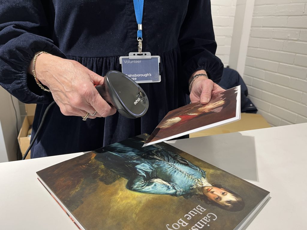 An image of a Gainsborough's House Shop Volunteer scanning a Gainsborough themed gift card.