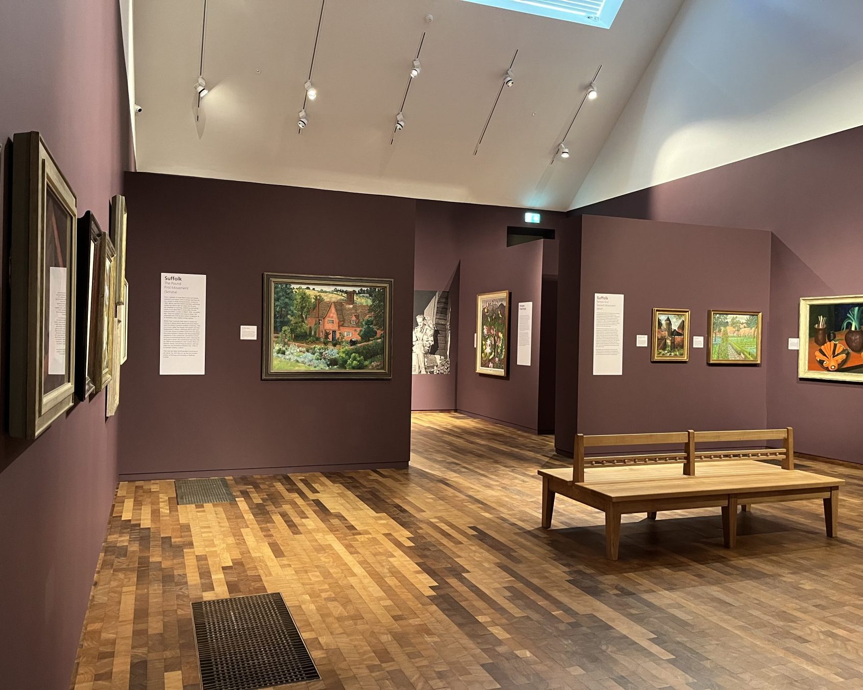 An image taken of the exhibition, Revealing Nature: The Art of Cedric Morris & Lett–Haines, in the Timothy & Mary Clode Exhibition Gallery at Gainsborough's House. There are various artworks hung on plum coloured walls.
