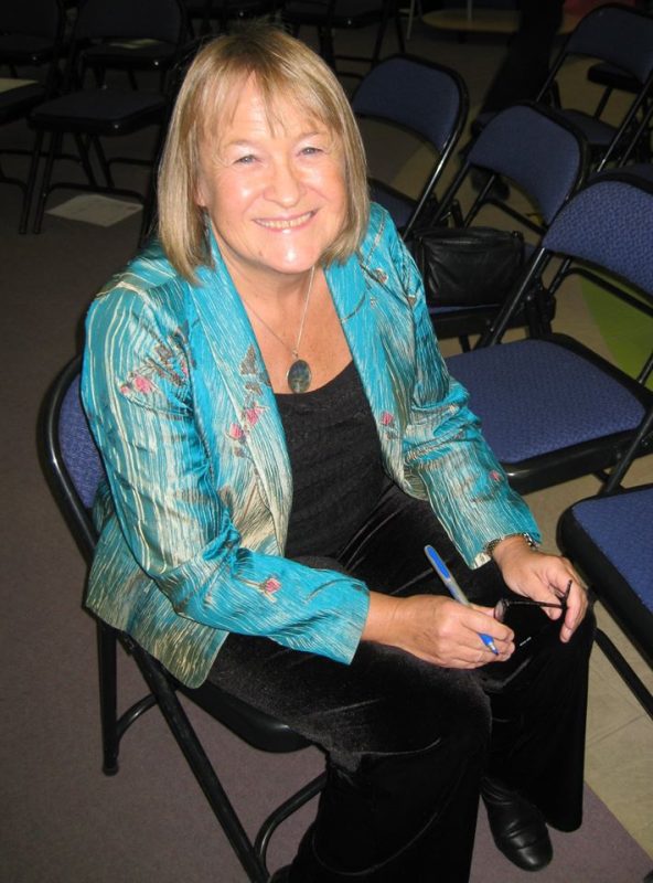 An image of Liz Trenow, a white woman with blonde hair. She is sat on a chair wearing a turqoise blazer. 