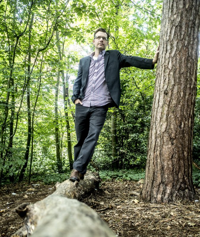 An image of Justin Hopper stood on a felled tree trunk in a forest, surrounded by trees. He is wearing a suit jacket and shirt. 