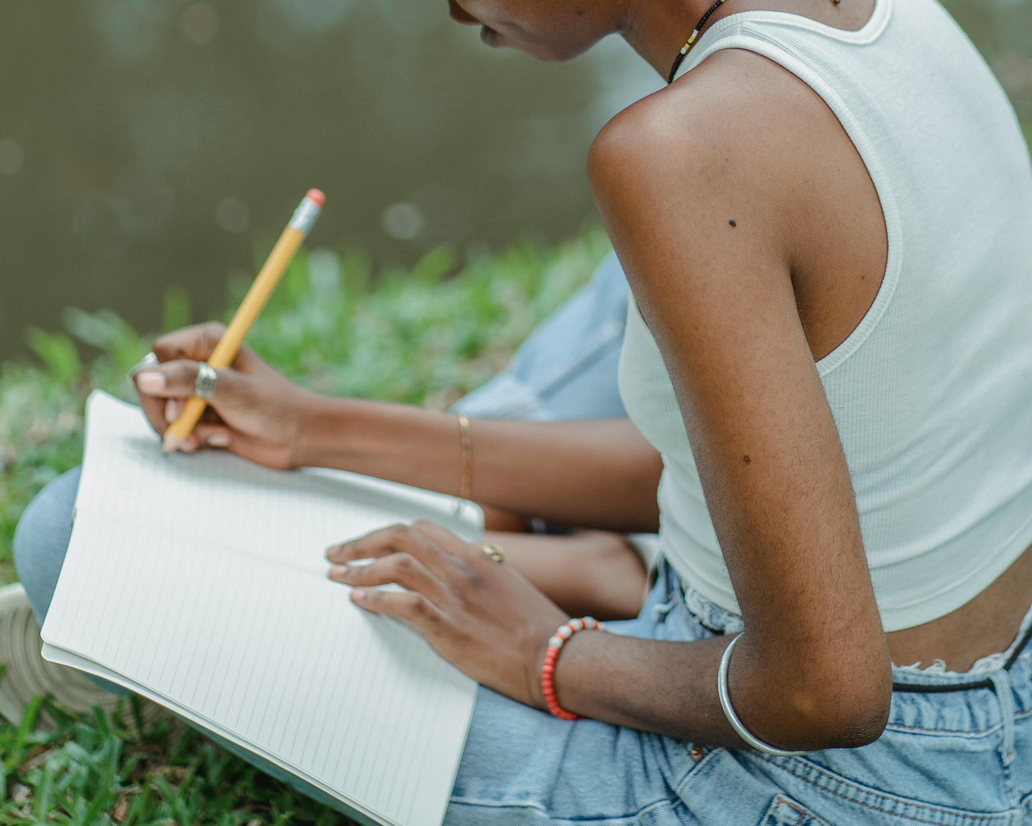 An image of a black person sat by a river bank with a notepad and pencil in hand as they write.