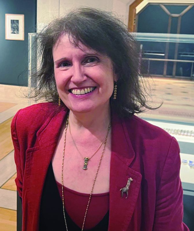 An image of Anne Desmet RA. A white woman with dark hair smiling to camera with a red jacket on. 