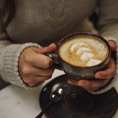 An image of a young woman in a grey jumper holding a cup of coffee.