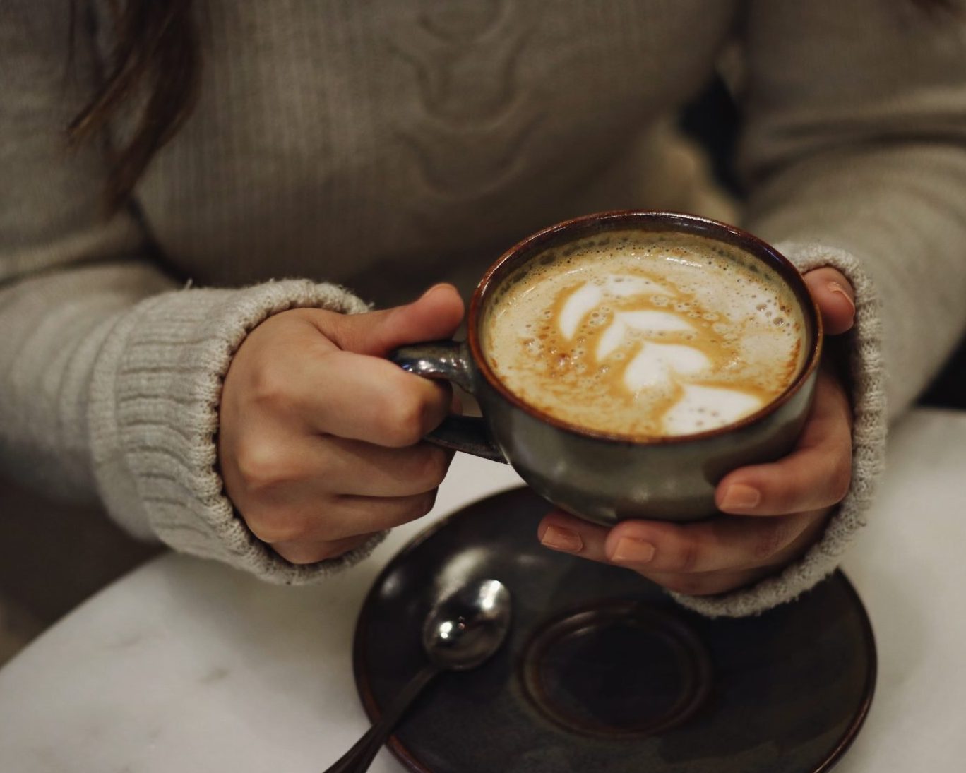 An image of a young woman in a grey jumper holding a cup of coffee.